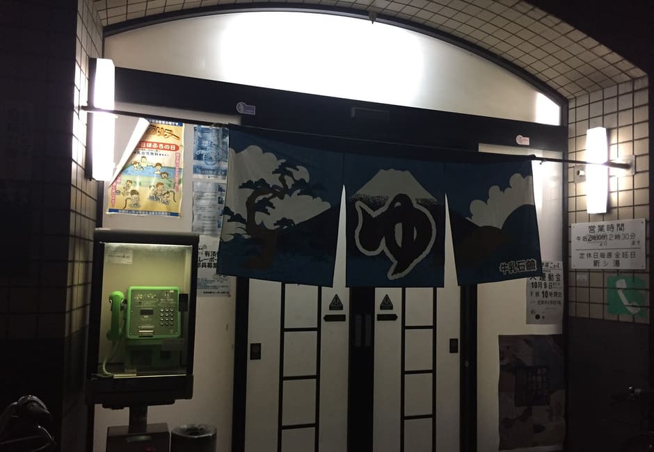 Entrance of Atarashi-yu sento at night, with its blue yu noren showing Mount Fuji, the hours board and a green public phone