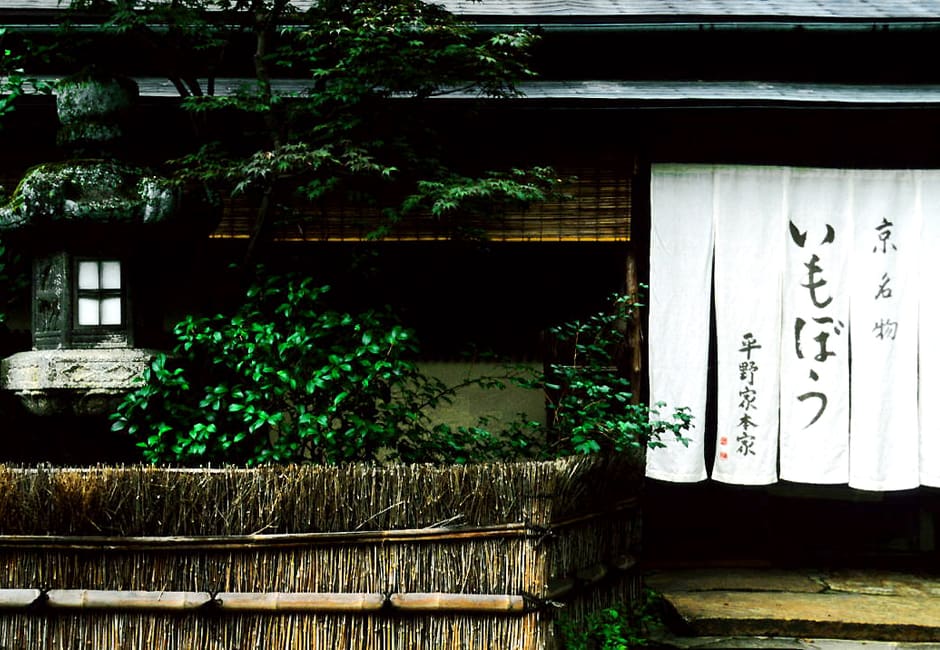 White noren reading imobo and Hiranoya Honke at the restaurant's entrance, beside a stone lantern and bamboo fence