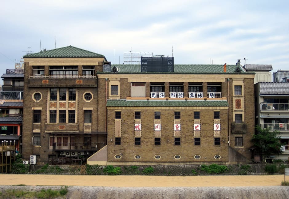 The brick Pontocho Kaburenjo theatre seen from the Kamo River bank, its green tiled roofs above the riverside path