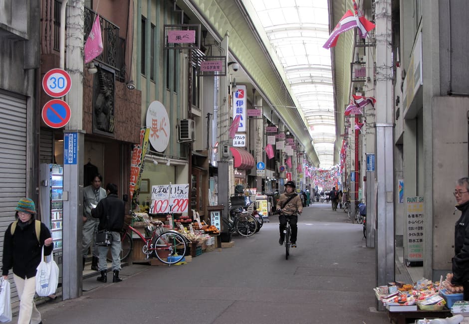 Inside the covered Sanjo-kai Shopping Street, with produce stalls, shop signs and a cyclist under the long glass roof